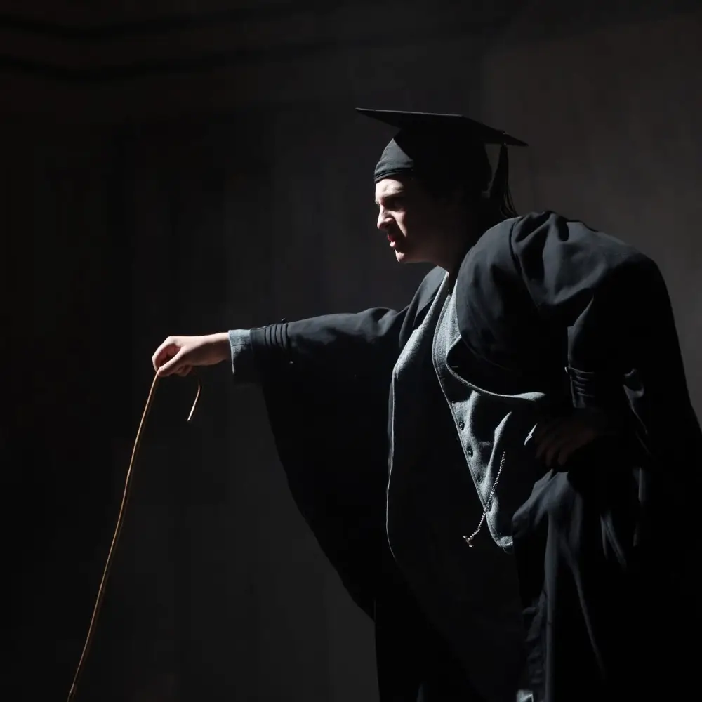 A performer in a graduation gown and mortarboard acting under dramatic stage lighting.
